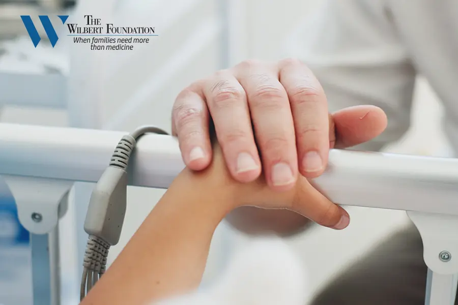 An adult hand holds a child’s hand on a hospital bed rail, with The Wilbert Foundation logo in the corner.