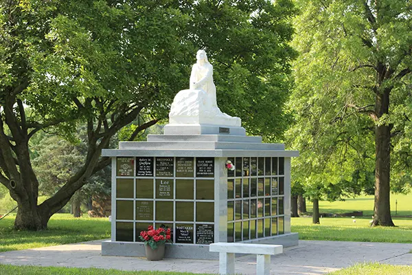 A stone columbarium in a park featuring a white sculpture of a kneeling figure on top and inscribed name plaques on the sides.