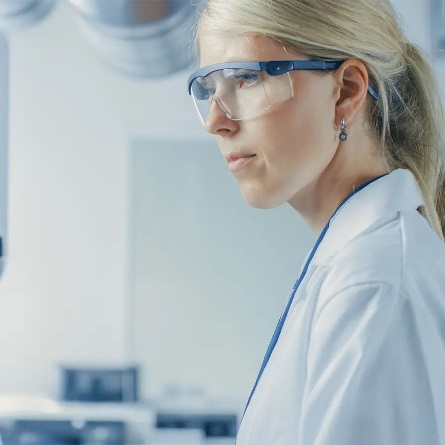 A scientist in a white lab coat and safety goggles looking toward the side.