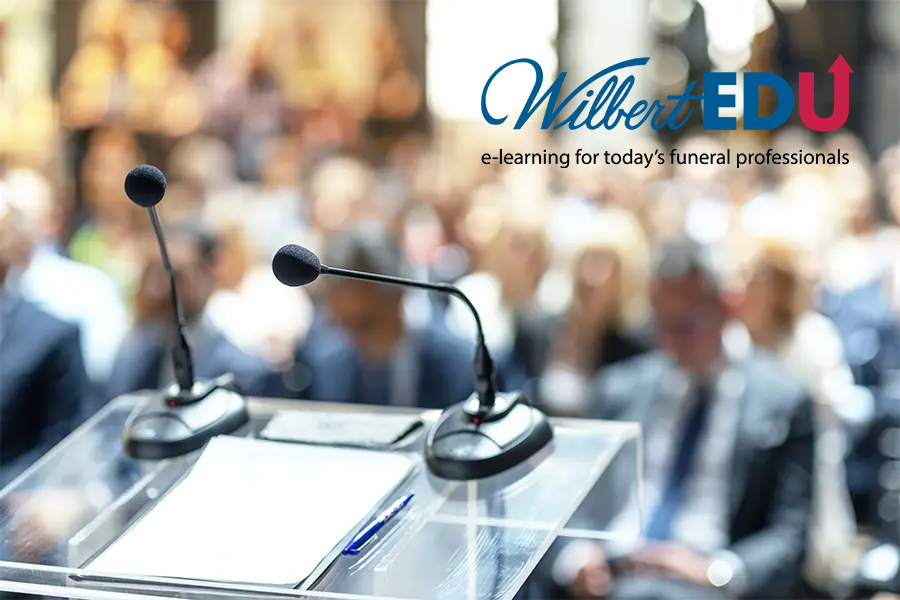 A transparent lectern with two microphones in front of a blurred audience, with the Wilbert EDU logo and tagline above.